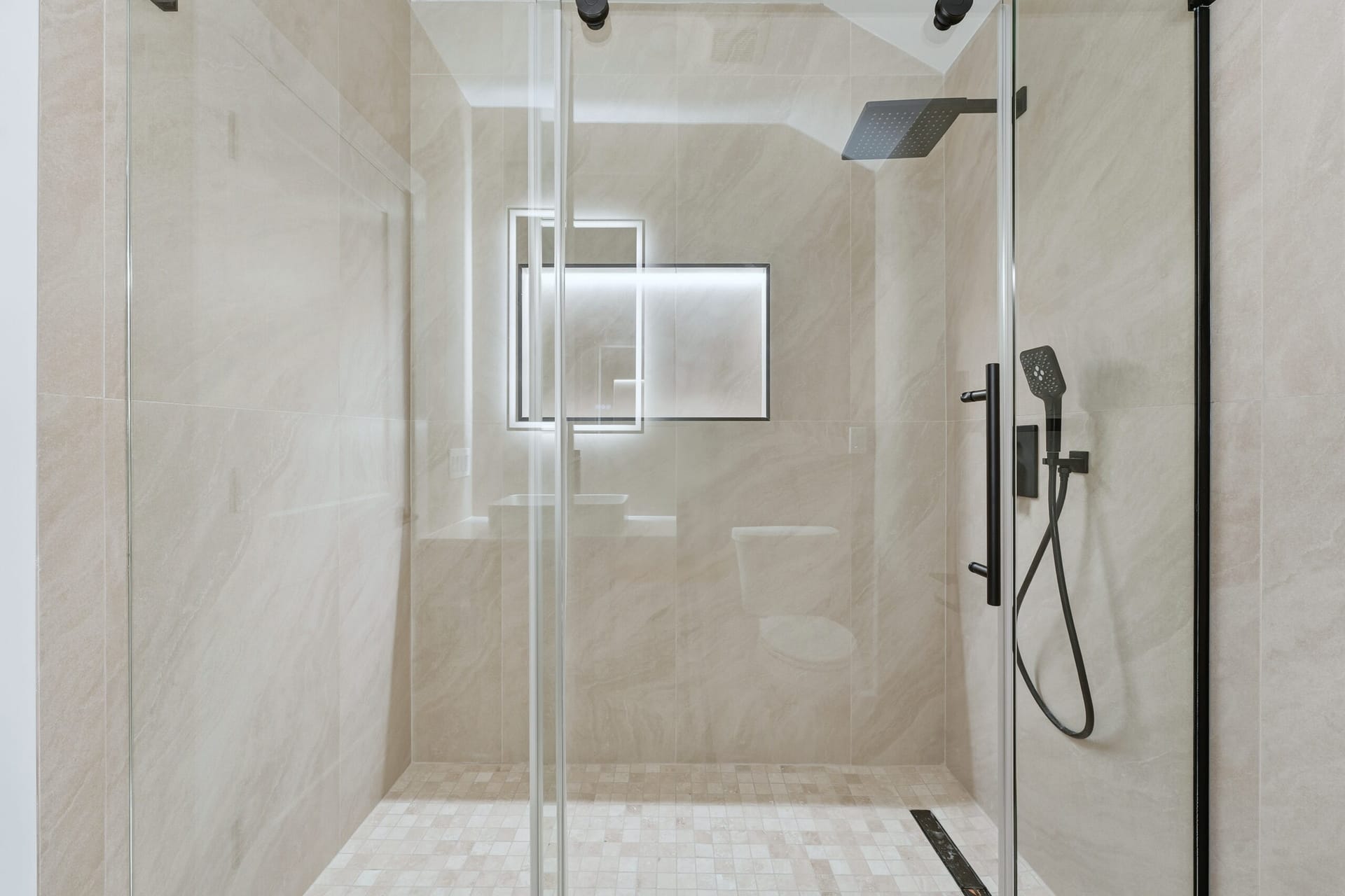 Walk-in shower clad in beige stone-look porcelain with dual matte-black rainfall heads and a mosaic floor